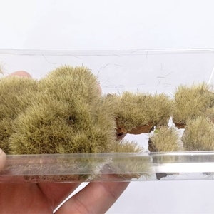 May include: A clear plastic container filled with small, green, fluffy tufts of artificial grass. The tufts are arranged in a variety of sizes and shapes.