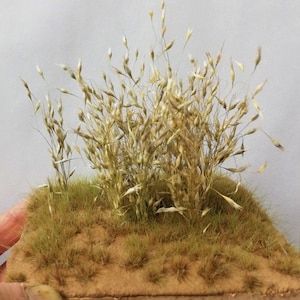 May include: A small patch of brown artificial grass with a cluster of tall, dried, light brown grass stalks in the center.