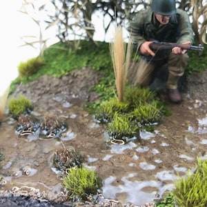 Puede incluir: Una figura de soldado en miniatura con uniforme militar, sosteniendo un rifle, está posicionada en un área cubierta de hierba con suelo fangoso. La figura está de pie cerca de un parche de hierba alta.