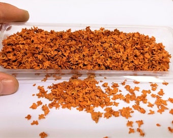 May include: A clear plastic container filled with small, orange, paper-like leaves. Some leaves have fallen out of the container onto a white surface.