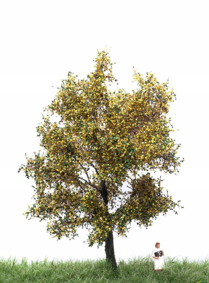 May include: A miniature model of a tree with green and yellow leaves. The tree is standing on a patch of green grass. A miniature figure of a person is standing next to the tree.