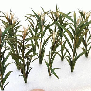 Puede incluir: Un grupo de 10 plantas verdes artificiales con flores blancas. Las plantas están dispuestas en fila sobre una superficie blanca.