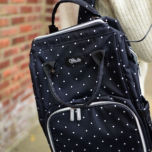 以下が含まれることがあります： 白のドット柄の黒いおむつバッグで、ジッパー付きのポケットが付いています。バッグにはトップハンドルと調節可能なショルダーストラップが付いています。バッグには「Mommy Chic」と書かれたラベルが付いています。