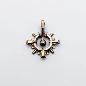 Könnte beinhalten: Ein detaillierter Anhänger im antiken Stil mit einem Sonnenstrahl-Design. Der Anhänger besteht aus einem metallischen Material mit goldener Oberfläche. Er hat eine zentrale Kugel, umgeben von einem kreisförmigen Rahmen und strahlenförmigen Elementen.