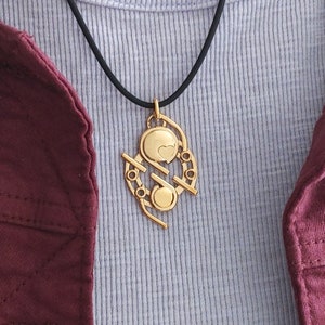 Puede incluir: Collar con colgante dorado y cordón negro. El colgante presenta un corazón y formas circulares con detalles lineales. El collar se muestra sobre una camiseta de canalé azul claro y una chaqueta burdeos.