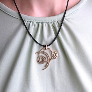 May include: A gold-colored pendant necklace with an abstract design. The pendant hangs from a black cord and features a series of interconnected swirls and circles. The necklace is displayed against a light green background.