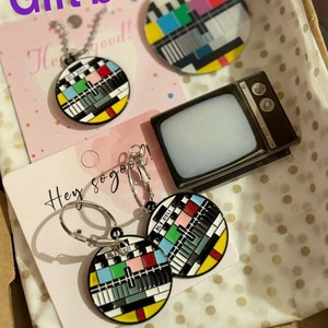 Könnte beinhalten: Geschenkbox mit einer Halskette, Ohrringen und einem kleinen Schlüsselanhänger in Form eines Fernsehers. Die Halskette und die Ohrringe haben einen runden Anhänger mit einem bunten Fernseh-Testmuster. Der Schlüsselanhänger ist schwarz und silber mit einem leeren Bildschirm.