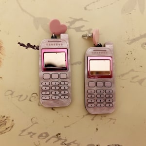 May include: A pair of pink cell phone earrings with a heart charm. The earrings are made of plastic and have a realistic cell phone design with a keypad and screen.