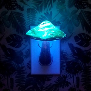 Pode incluir: Uma luz noturna em forma de cogumelo verde conectada a uma tomada el&eacute;trica. A luz est&aacute; acesa e emite um brilho azul.