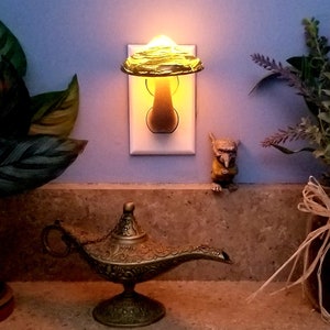 Pode incluir: Uma luz noturna em forma de cogumelo de vidro verde conectada a uma tomada branca. A luz noturna est&aacute; acesa e projeta um brilho quente. Uma pequena l&acirc;mpada de g&ecirc;nio dourada est&aacute; em um balc&atilde;o em primeiro plano.