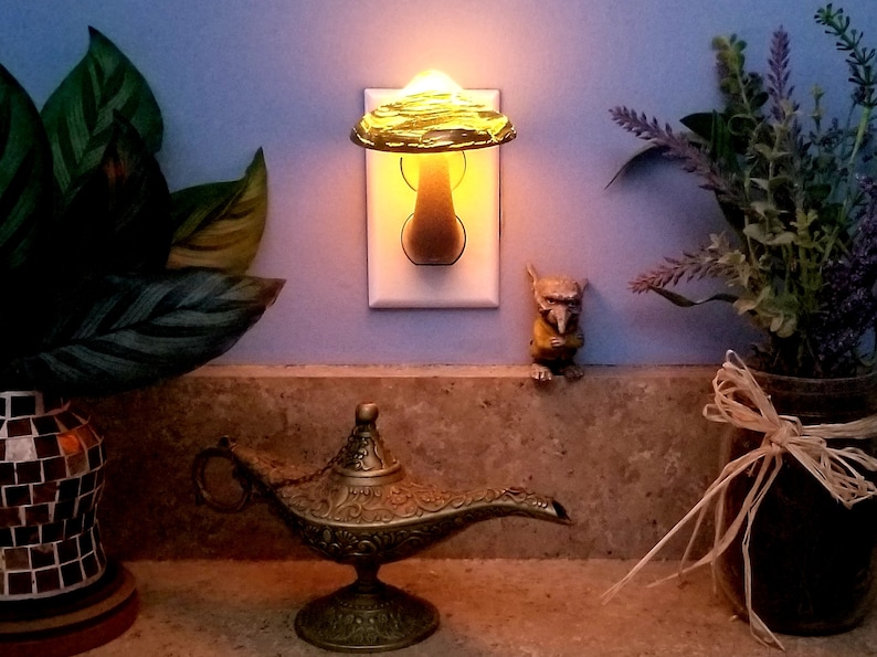 Pode incluir: Uma luz noturna em forma de cogumelo de vidro verde conectada a uma tomada branca. A luz noturna est&aacute; acesa e projeta um brilho quente. Uma pequena l&acirc;mpada de g&ecirc;nio dourada est&aacute; em um balc&atilde;o em primeiro plano.