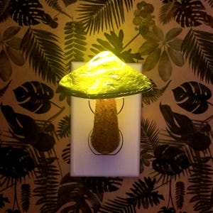 Pode incluir: Uma luz noturna em forma de cogumelo de vidro amarelo conectada a uma tomada el&eacute;trica. A luz noturna est&aacute; em uma parede com um padr&atilde;o de folhas tropicais.