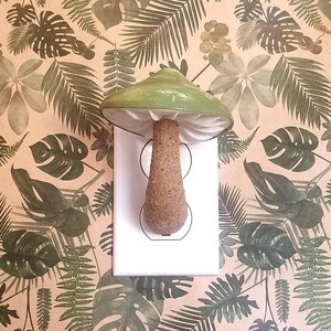 Pode incluir: Uma tampa de interruptor de luz em forma de cogumelo de cer&acirc;mica verde com um caule castanho. A tampa est&aacute; montada numa tomada el&eacute;trica branca. O fundo &eacute; um papel de parede bege com um padr&atilde;o de folhas tropicais verdes.