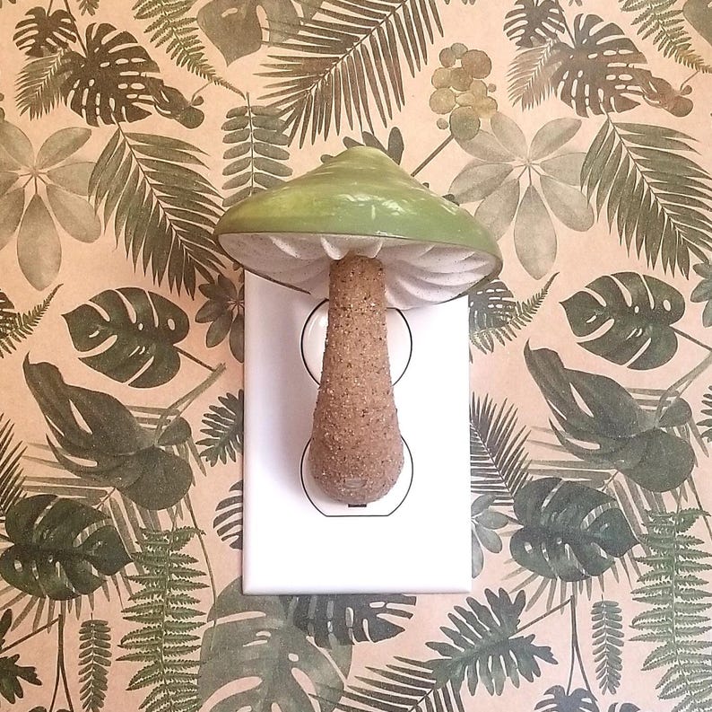 Pode incluir: Uma tampa de interruptor de luz em forma de cogumelo de cer&acirc;mica verde com um caule castanho. A tampa est&aacute; montada numa tomada el&eacute;trica branca. O fundo &eacute; um papel de parede bege com um padr&atilde;o de folhas tropicais verdes.