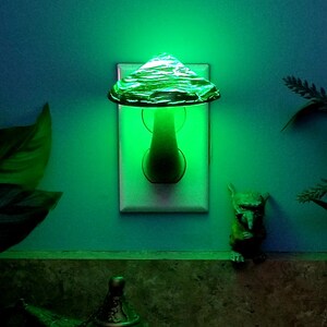 Pode incluir: Uma luz noturna em forma de cogumelo de vidro verde, conectada a uma tomada el&eacute;trica. A luz noturna brilha em verde.