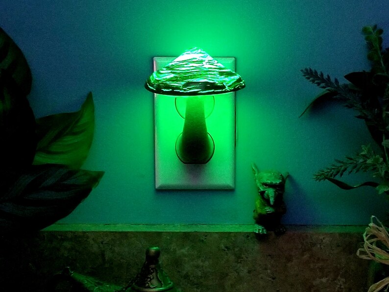 Pode incluir: Uma luz noturna em forma de cogumelo de vidro verde, conectada a uma tomada el&eacute;trica. A luz noturna brilha em verde.