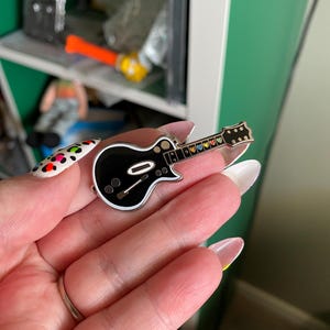 Könnte beinhalten: Eine schwarze und silberne Gitarrennadel mit weißem Rand und bunten Herzen auf dem Griffbrett.