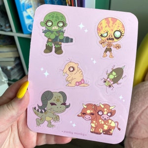 May include: A sheet of pink stickers featuring cartoon monsters. The stickers include a green monster with a gun, a pink monster with a bow, a brown monster with two heads, a brown beetle, and a yellow monster with a skull.