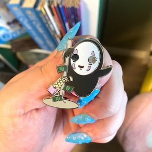 May include: A gold-colored enamel pin featuring a cartoon character from the movie Spirited Away. The character is a white, faceless spirit with black hair and a pink and blue face. The character is holding a pole with money wrapped around it. The pin is on a hand with blue nail polish.
