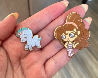 May include: Two enamel pins featuring characters from the Disney movie Hercules. The first pin is a light blue Pegasus with a light blue mane and tail. The second pin is a brown-haired woman with a pink dress and a gold crown.