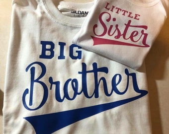 May include: Two white t-shirts with blue and pink text. The larger shirt says "BIG BROTHER" in blue with a blue baseball-style arc underneath. The smaller shirt says "LITTLE SISTER" in pink with a pink baseball-style arc underneath.