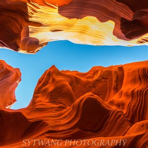 May include: A photograph of a sandstone canyon with layered rock formations in shades of orange and red. The sky is a bright blue, visible between the canyon walls. The image is labeled "SYTWANG PHOTOGRAPHY".