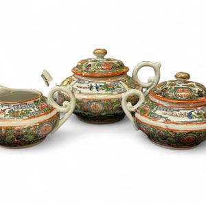 May include: A porcelain tea set with a teapot, sugar bowl, and creamer. The set features a detailed, colorful design with floral and landscape motifs, accented with gold trim. The handles are curved and the lids have gold finials.