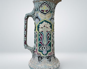 May include: A tall, ceramic pitcher with a wide, flared mouth and a handle. The pitcher is decorated with a colorful, intricate floral pattern in shades of green, purple, and yellow. The pattern includes stylized flowers, leaves, and geometric designs.