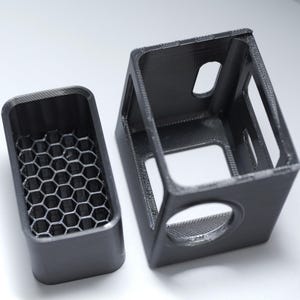 May include: Two black 3D-printed rectangular cases. One case has a honeycomb pattern inside. The other case is open with rectangular and circular openings. The cases are likely for electronics or small objects.