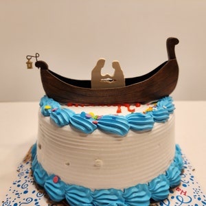 May include: A round white cake decorated with blue frosting swirls and colorful sprinkles. A wooden boat cake topper with two figures is placed on top. A small lantern hangs from the boat.