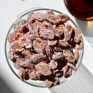 May include: A close-up of a bowl filled with dried apricot halves. The apricots are a light brown color and have a slightly powdery texture. The bowl is clear glass and is sitting on a white surface.