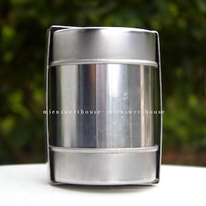 May include: A stainless steel container with a cylindrical body and a lid. The container is held in place by a metal frame. The text "miensweethouse" is printed on the side. The background is a blurred green.