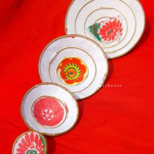 May include: Four white, circular decorative pieces with floral designs in red, green, and yellow, arranged on a red background. Each piece has a gold-colored rim and a spiral design. The sizes vary, with the largest at the top.