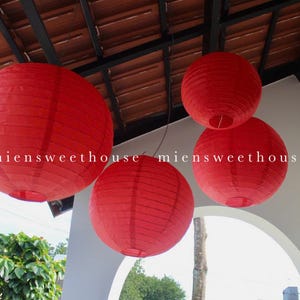 May include: Four red paper lanterns of varying sizes hang from a ceiling. The lanterns are round with a ribbed texture. The text "miensweethouse" is visible on the lanterns. The lanterns are suspended in front of a white wall and a brown roof.