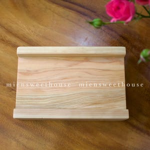 Puede incluir: Un soporte de teléfono de madera de color claro con el texto "miensweethouse" en blanco. El soporte tiene forma rectangular con bordes elevados. Hay rosas rosas en el fondo.