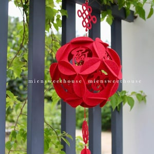 May include: Red felt Chinese lantern with intricate cutouts and a knotted tassel hanging from a fence. The lantern has Chinese characters on it.