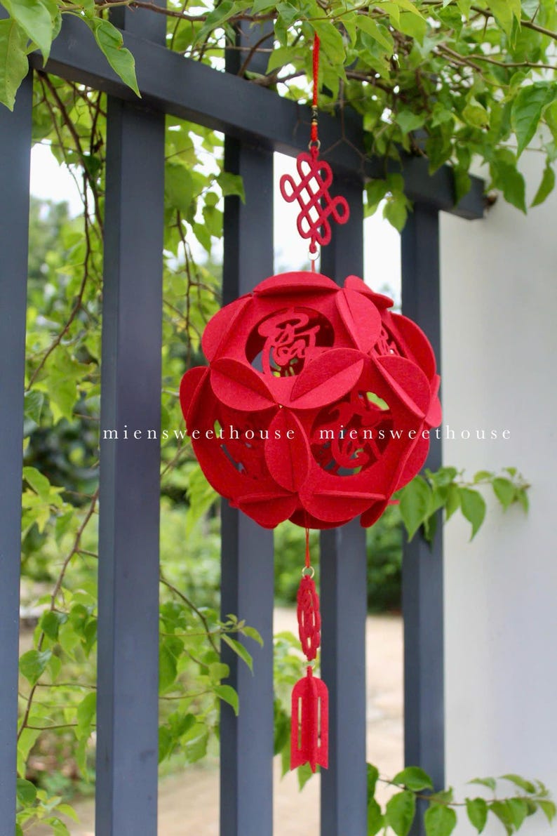 May include: Red felt Chinese lantern with intricate cutouts and a knotted tassel hanging from a fence. The lantern has Chinese characters on it.