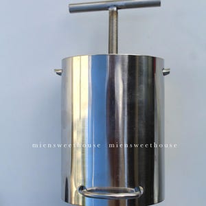 Könnte beinhalten: Eine Edelstahl-Lebensmittelpresse mit einem T-förmigen Griff. Die Presse hat einen zylindrischen Körper mit einem Griff an der Seite.