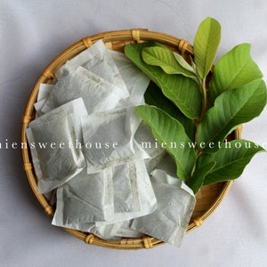 以下が含まれることがあります： 小さな正方形のティーバッグと新鮮な緑の葉が入った丸い柳のバスケット。 ティーバッグは白く、葉は鮮やかな緑色です。 画像には「miensweethouse」の文字が印刷されています。