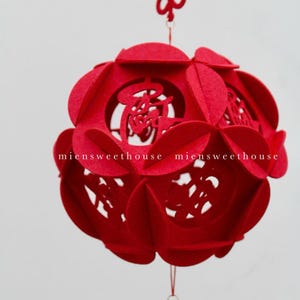 May include: A red felt Chinese lantern with intricate cutouts and a tassel hanging from the bottom. The lantern is decorated with Chinese characters and a red knot at the top.