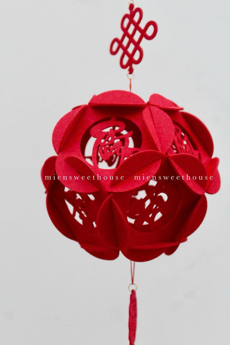 May include: A red felt Chinese lantern with intricate cutouts and a tassel hanging from the bottom. The lantern is decorated with Chinese characters and a red knot at the top.
