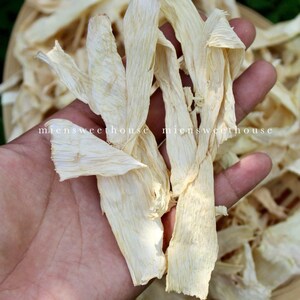 May include: A hand holding a bunch of dried, white, thin, and flat pieces of bamboo shoots. The bamboo shoots are arranged in a fan shape. The background is a basket filled with more bamboo shoots.