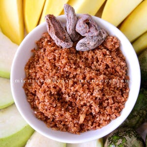 Pode incluir: Uma tigela branca cheia de uma mistura marrom e granulada, coberta com frutas secas. A tigela é cercada por fatias de frutas verdes e uma pequena fruta redonda verde.