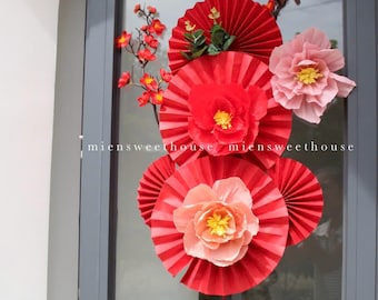 May include: Red and pink paper flower decorations with a red tassel hanging from the bottom. The flowers are arranged in a circle on a glass door.