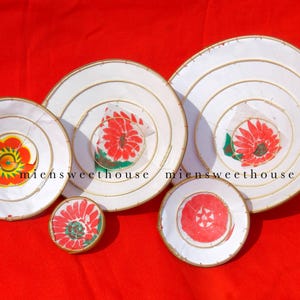 May include: A collection of white, circular decorative items with gold trim and floral designs in red, yellow, and green. The items are arranged on a red background. The text "miensweethouse" is visible.