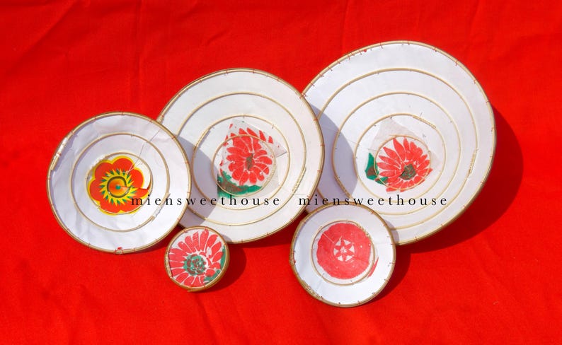 May include: A collection of white, circular decorative items with gold trim and floral designs in red, yellow, and green. The items are arranged on a red background. The text "miensweethouse" is visible.