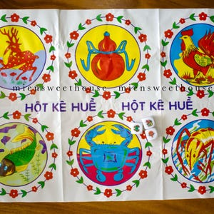 Puede incluir: Un colorido tablero de juego vietnamita con seis ilustraciones circulares de animales y objetos, incluyendo un ciervo, un gallo y un cangrejo, cada uno rodeado por una corona floral. El texto "HỘT KÊ HUẾ" está impreso en el tablero, con tres dados.