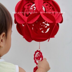 May include: A red felt Chinese lantern with intricate cutouts and a tassel hanging from the bottom. The lantern is decorated with Chinese characters. A child's hand reaches out to touch the lantern.