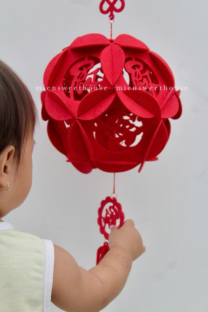 May include: A red felt Chinese lantern with intricate cutouts and a tassel hanging from the bottom. The lantern is decorated with Chinese characters. A child's hand reaches out to touch the lantern.