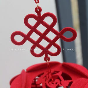 May include: A red felt Chinese knot, also known as a lucky knot, hangs from a red string. The knot is a symbol of good fortune and is often used in Chinese decorations. The knot is attached to a red felt ball with cutouts.